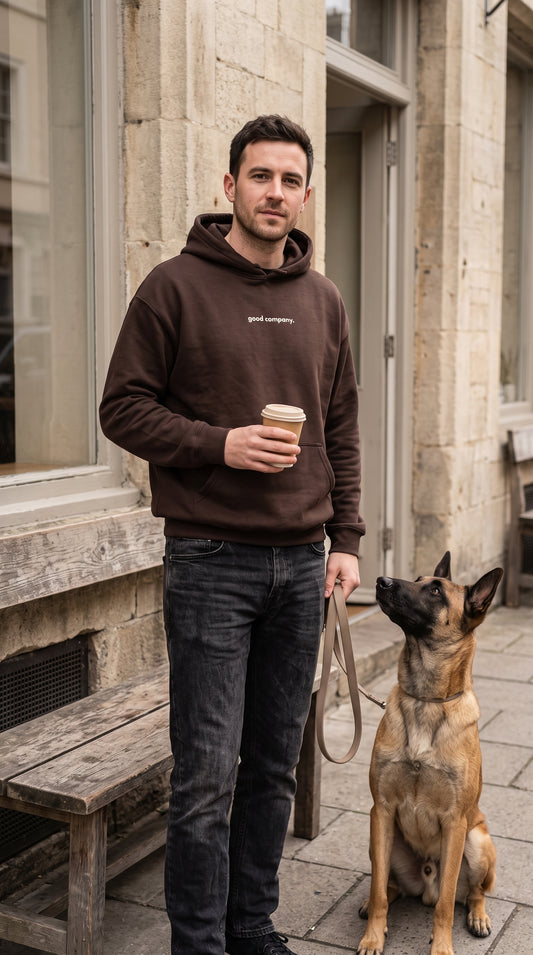 Man wearing a brown hoodie with cream “good company.” chest lettering, holding coffee beside a Belgian Malinois outside a café.