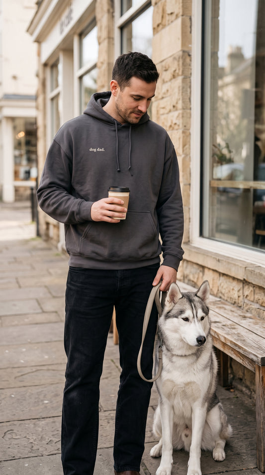 Man wearing a charcoal hoodie with small cream “dog dad.” lettering, holding coffee beside a husky outside a café.