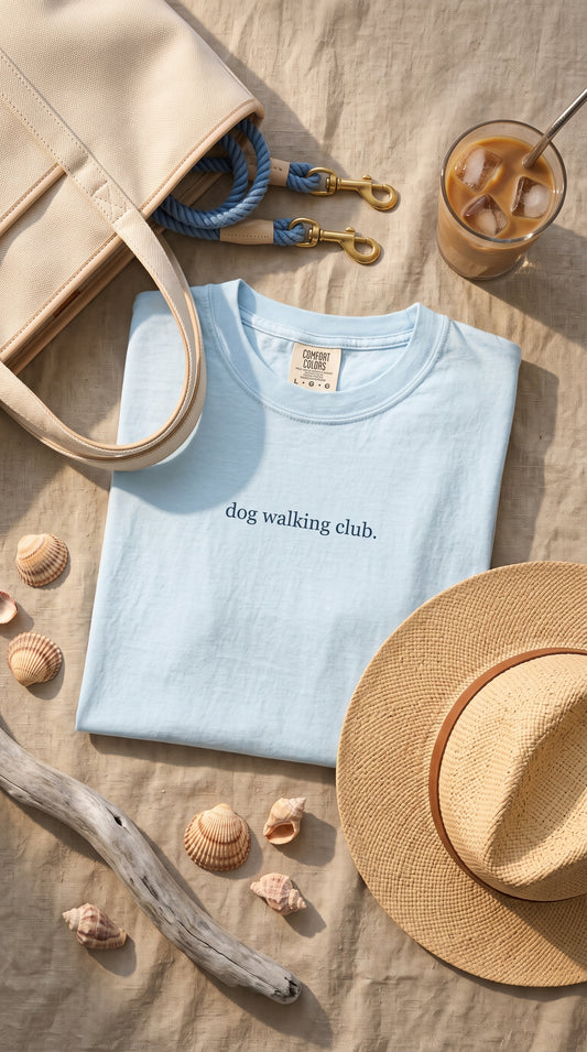 A light blue crewneck t-shirt with 'dog walking club.' printed in black across the chest, styled in a flat lay on a beige linen surface with a straw hat, seashells, driftwood, a tote bag, and an iced coffee.