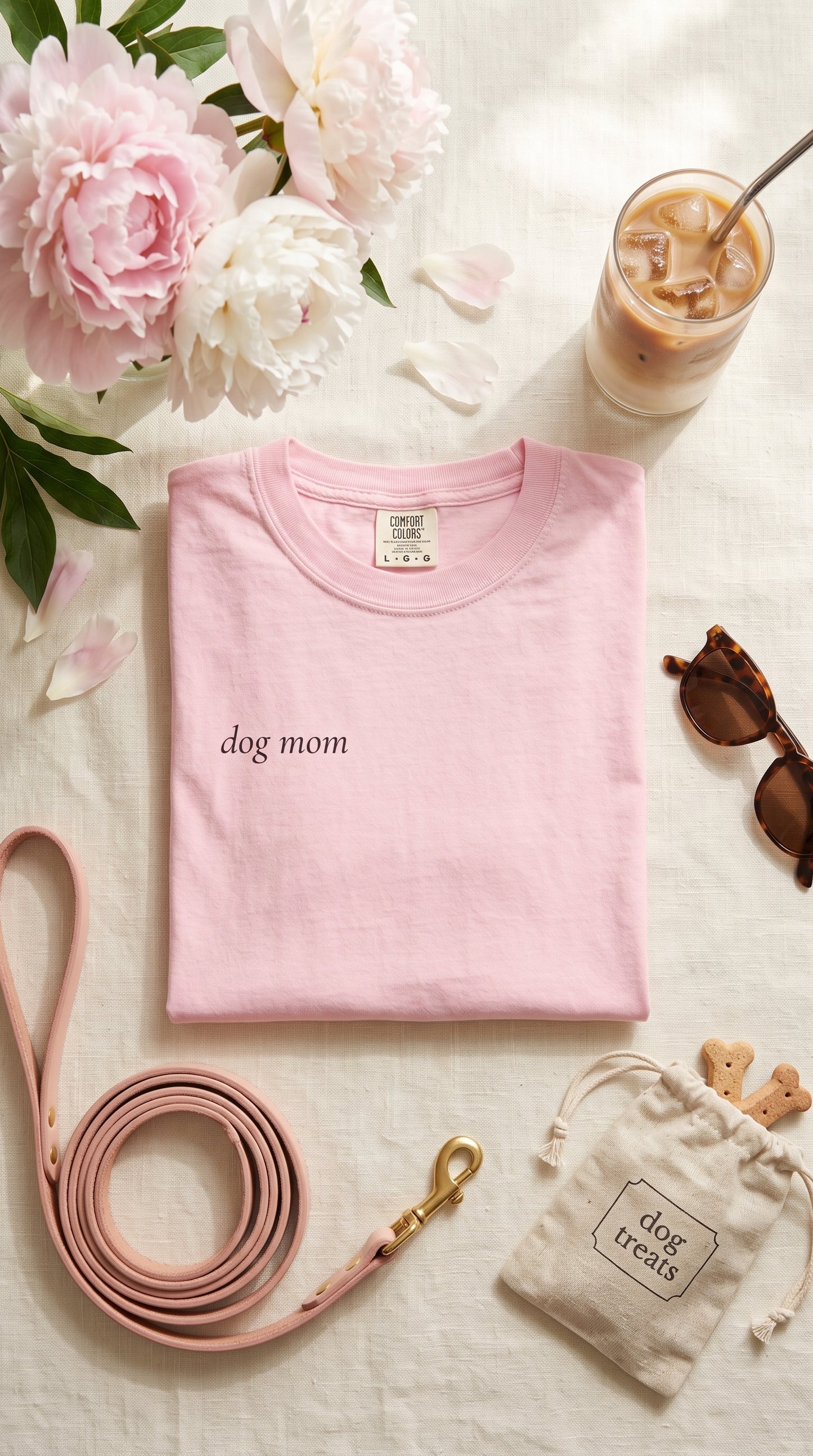 A folded light pink T-shirt with 'dog mom' printed in black script on the chest, styled in a flat lay with a pink leash, sunglasses, a drawstring bag, and flowers on a light fabric background.