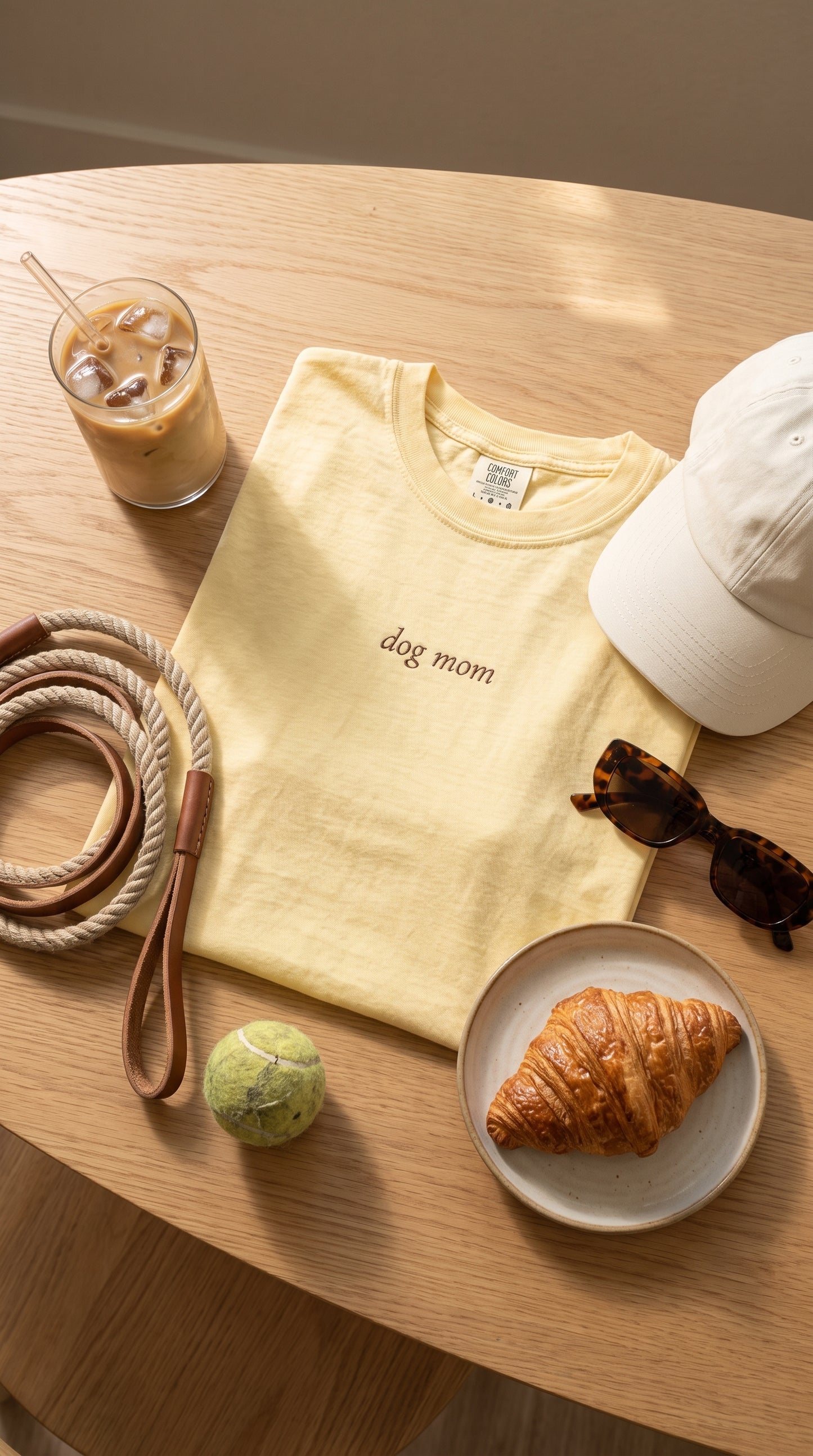 A pale yellow crewneck t-shirt with 'dog mom' embroidered in black on the chest, styled in a flat-lay on a light wood table with a white baseball cap, sunglasses, an iced coffee, a croissant on a plate, and a rope bag.