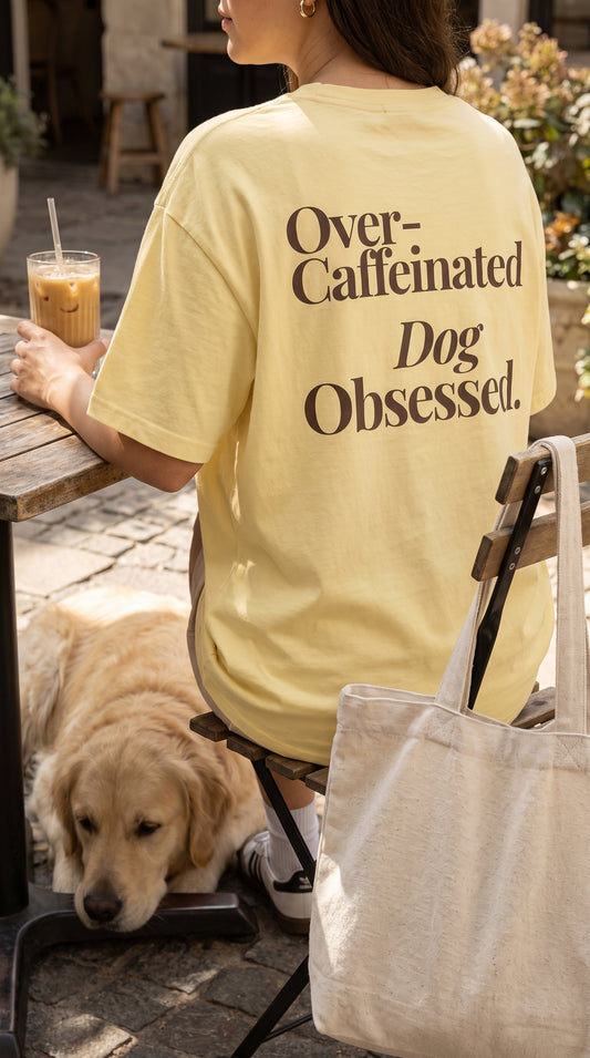 A woman sitting at an outdoor cafe table wearing a pale yellow t-shirt with the text 'Over-Caffeinated Dog Obsessed.' printed in dark brown on the back.