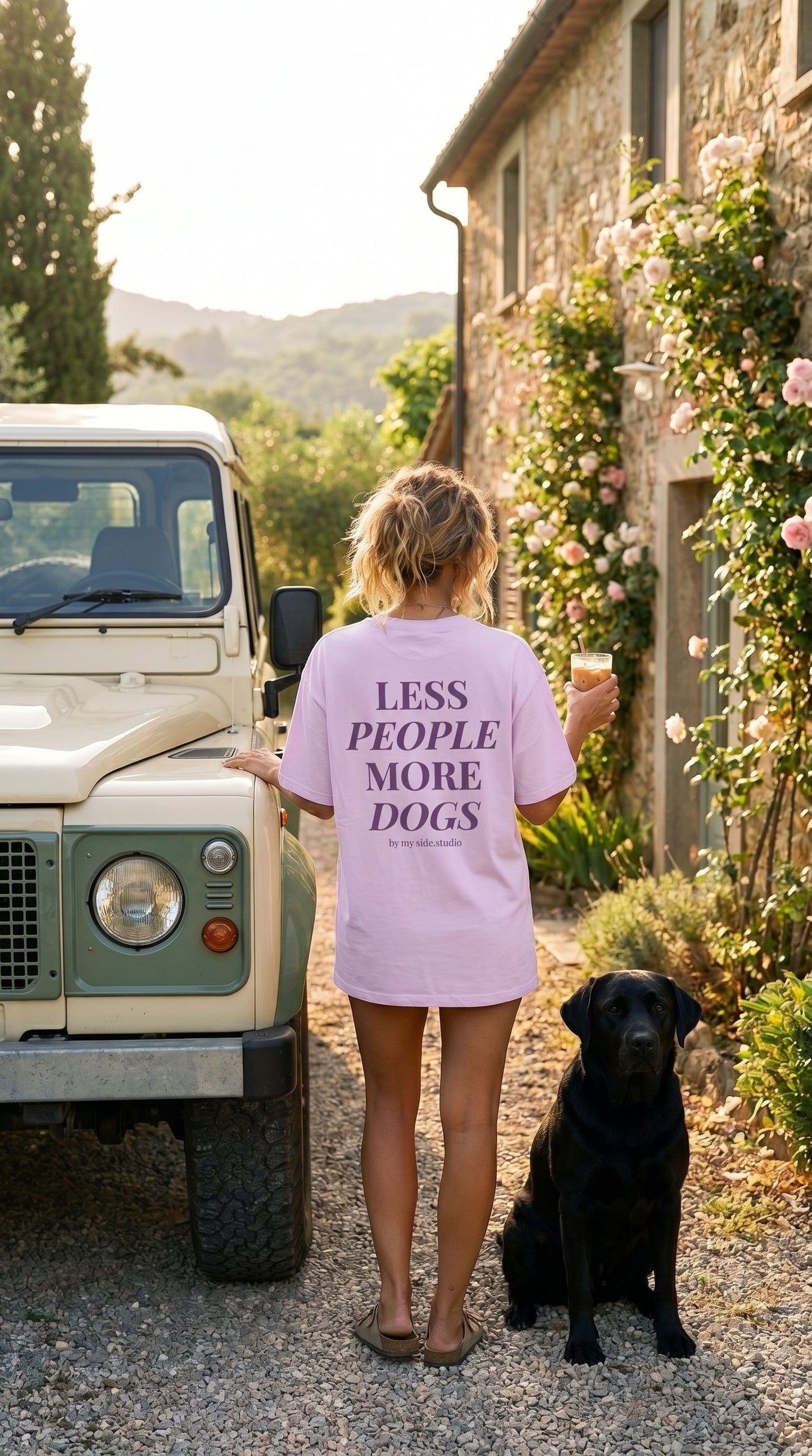Person wearing a shirt with 'LESS PEOPLE MORE DOGS' text standing next to a black dog and a vintage car.