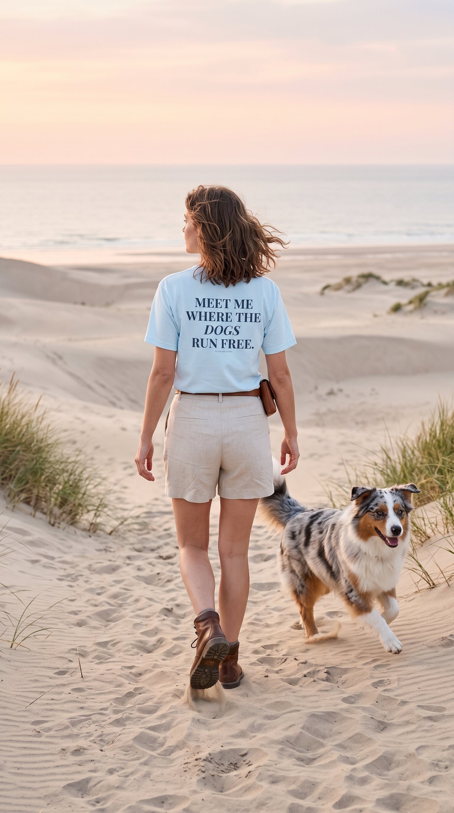 A woman walking on sandy dunes with her dog, seen from behind, wearing a light blue graphic T-shirt that reads 'MEET ME WHERE THE DOGS RUN FREE'.