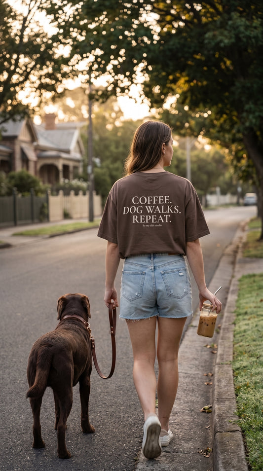 Cinematic lifestyle photograph of a woman walking through a quiet residential neighborhood at sunrise wearing an oversized espresso brown Comfort Colors t-shirt. The back graphic reading "Coffee. Dog Walks. Repeat." is fully visible. She carries an iced coffee while walking a Chocolate Labrador beside tree-lined streets. Warm golden hour light, subtle motion blur, luxury clothing campaign, premium editorial photography, calm slow-living aesthetic, muted color palette, shallow depth of field.