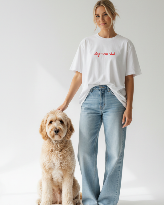 Woman wearing a white t-shirt with 'dog mom club' text and light blue jeans, standing next to a dog on a plain background.