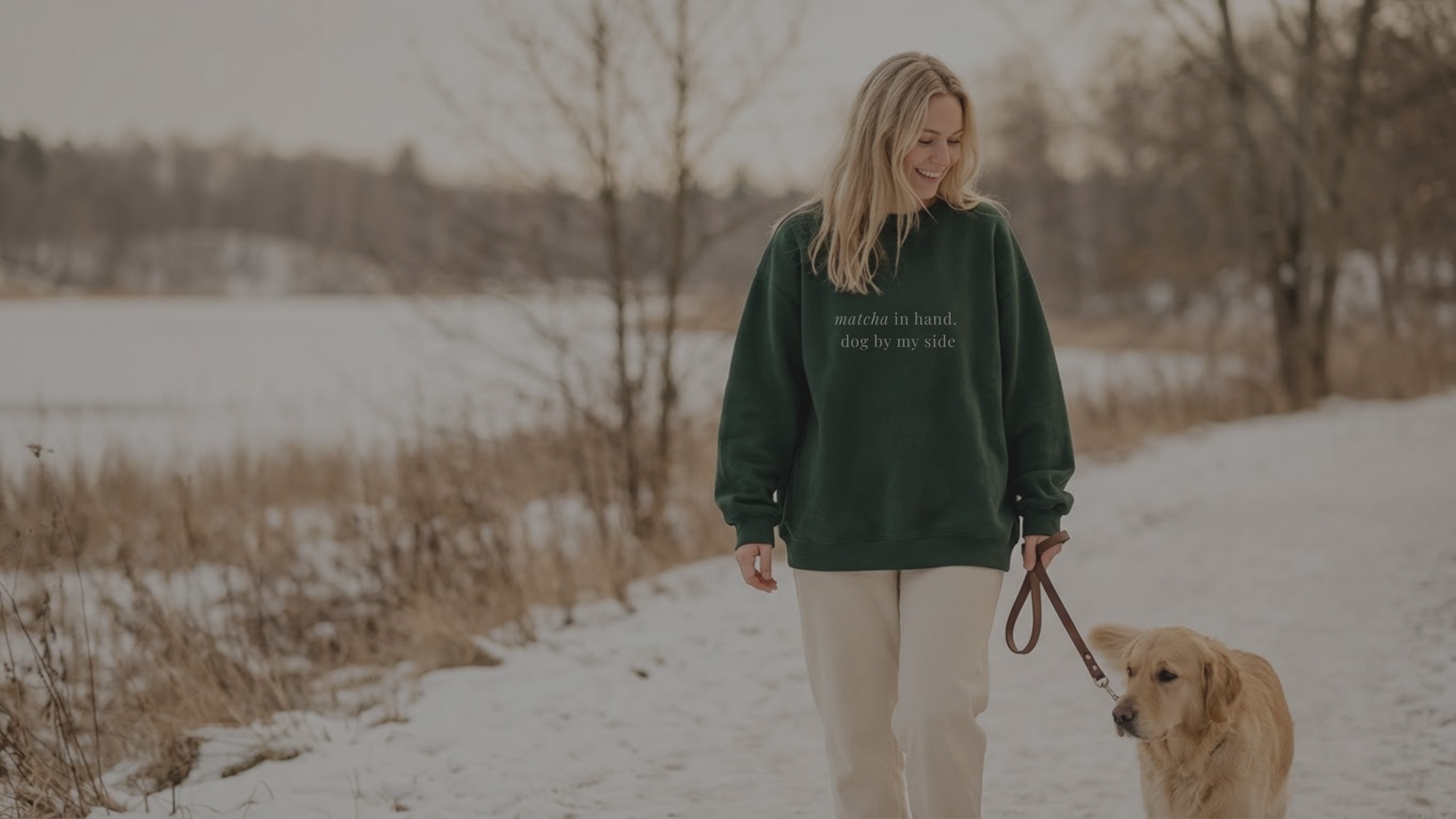 Woman walking a dog on a snowy path with a body of water and trees in the background