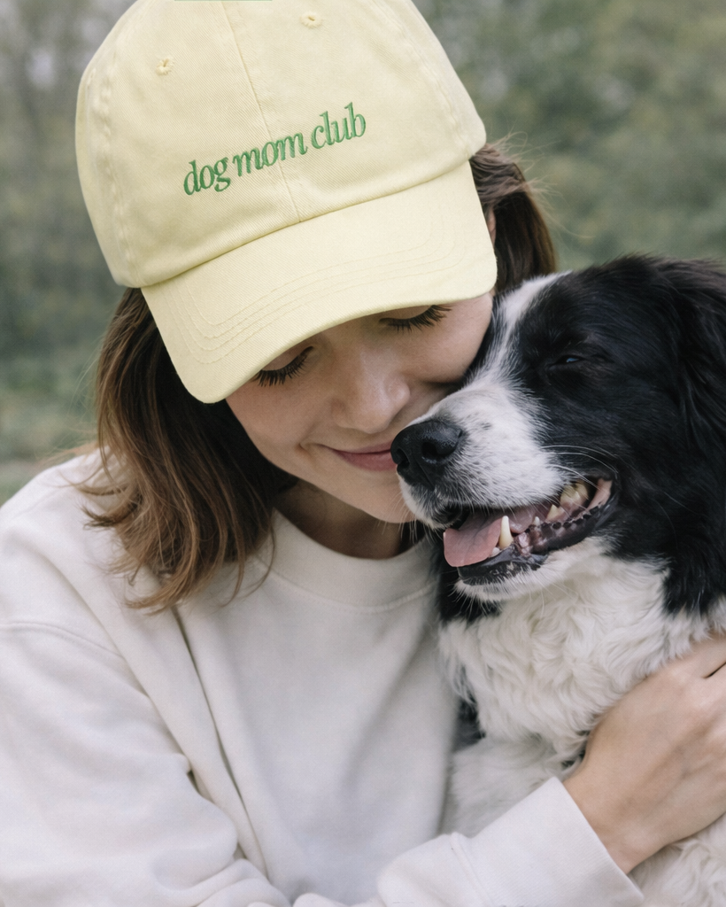 Woman wearing a 'dog mom club' cap hugging a black and white dog outdoors.