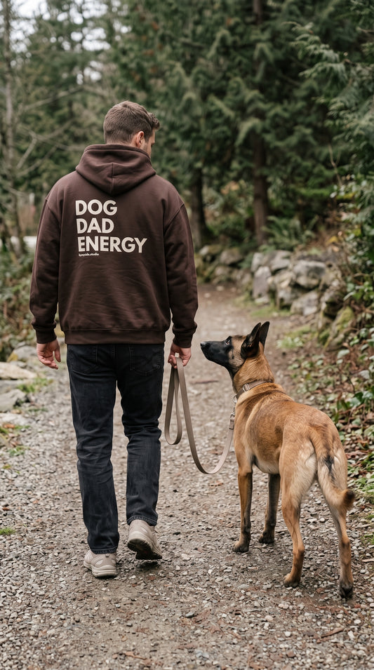 Man wearing a brown “Dog Dad Energy” hoodie, viewed from behind, walking a Belgian Malinois along a gravel trail.