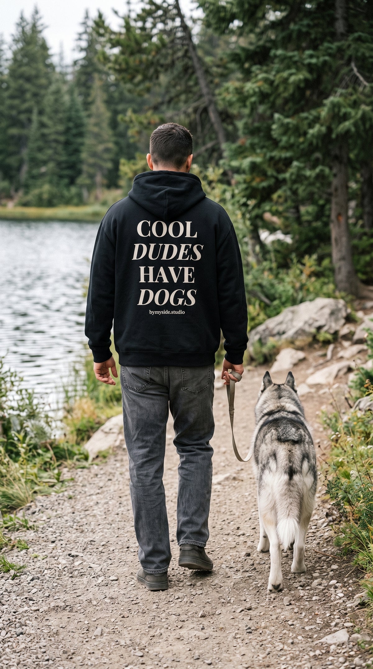 Back view of a man wearing a black “Cool Dudes Have Dogs” hoodie with cream lettering, walking a husky beside a lake.