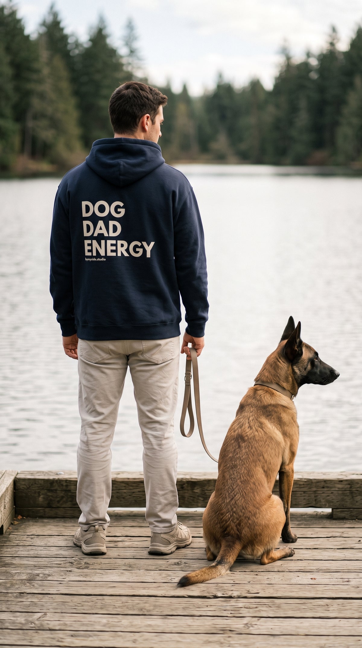 Back view of a man wearing a navy “Dog Dad Energy” hoodie with cream lettering, standing beside a Belgian Malinois on a lakeside dock.