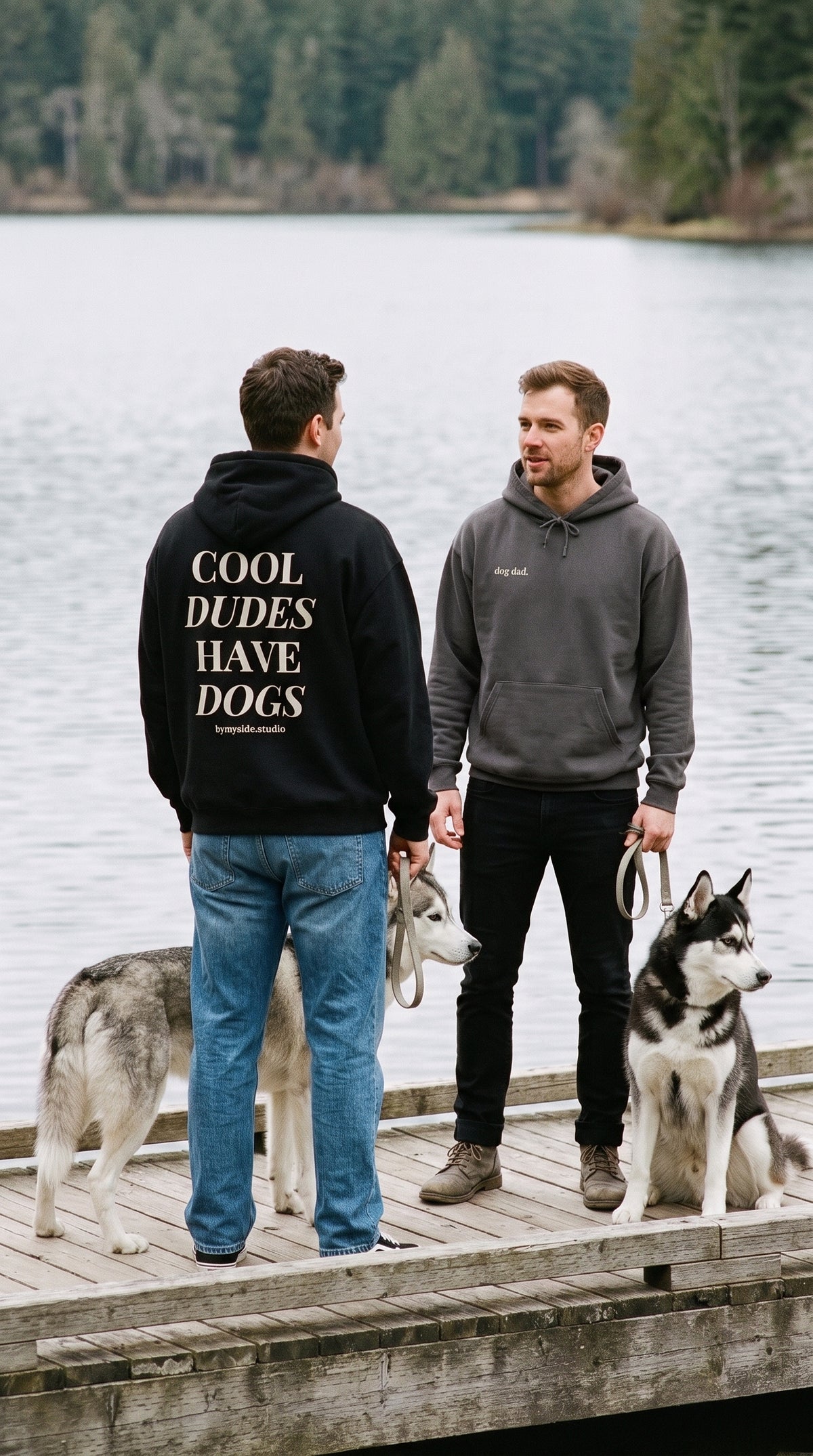 Two men with huskies on a lakeside dock, showing the black hoodie’s “Cool Dudes Have Dogs” back and charcoal hoodie’s “dog dad.” front.