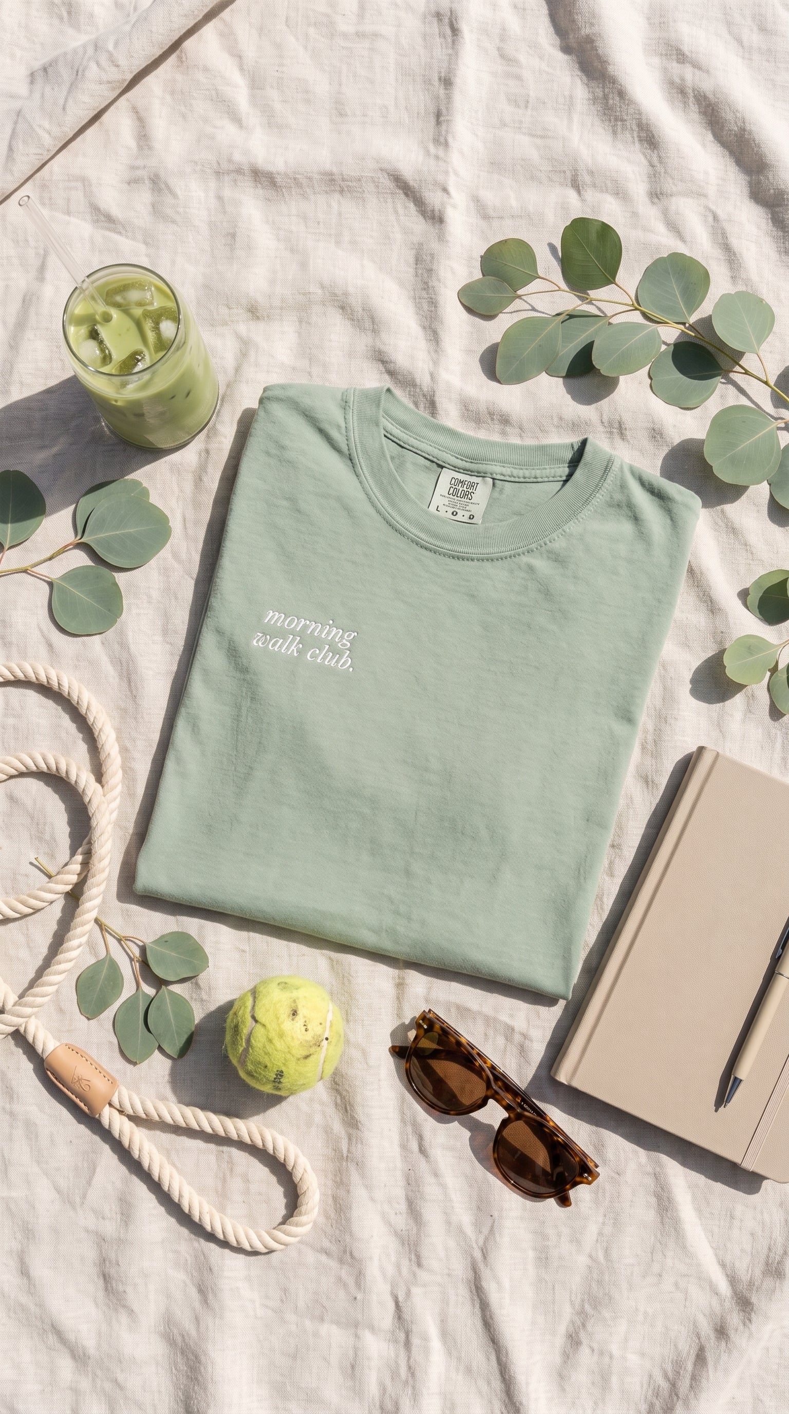 A folded sage green t-shirt with white embroidered text reading 'morning walk club' is displayed in a styled flat-lay arrangement on a textured white linen background, surrounded by eucalyptus leaves, sunglasses, a notebook, a rope, and a drink.
