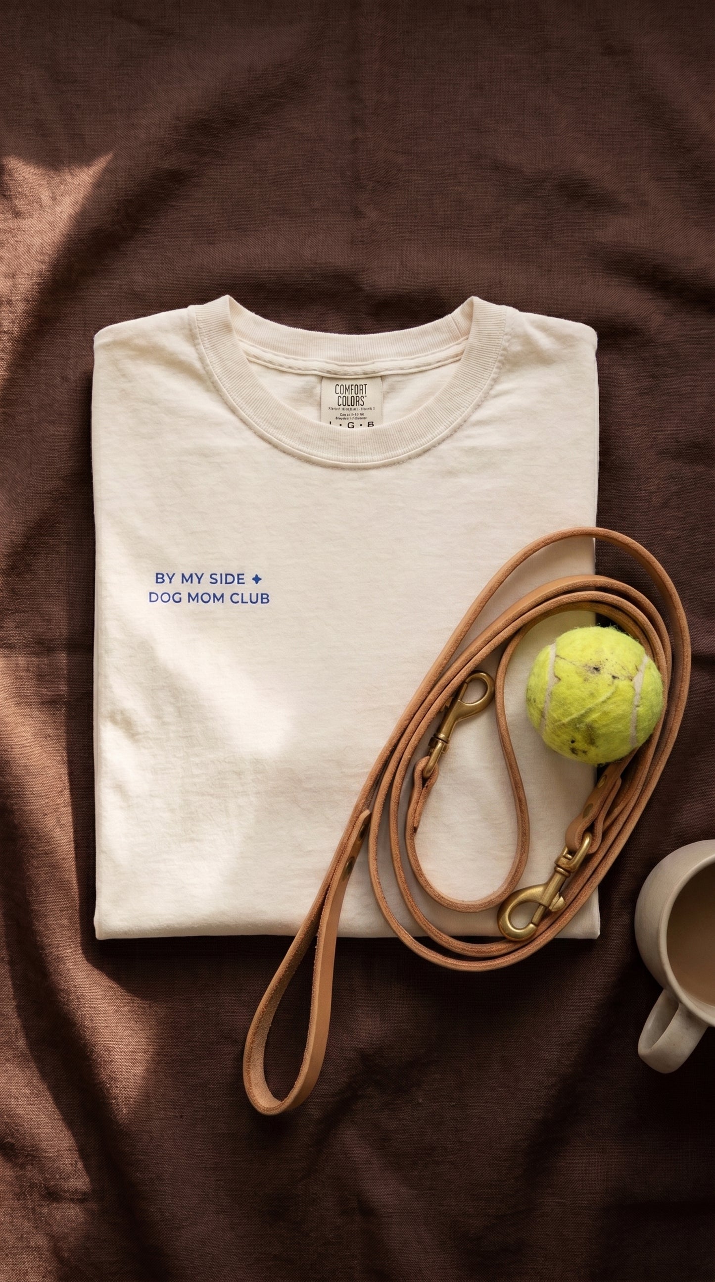 A folded off-white t-shirt with blue text reading 'BY MY SIDE + DOG MOM CLUB' is styled on a brown fabric background with a tan dog leash, a yellow tennis ball, and a cup.