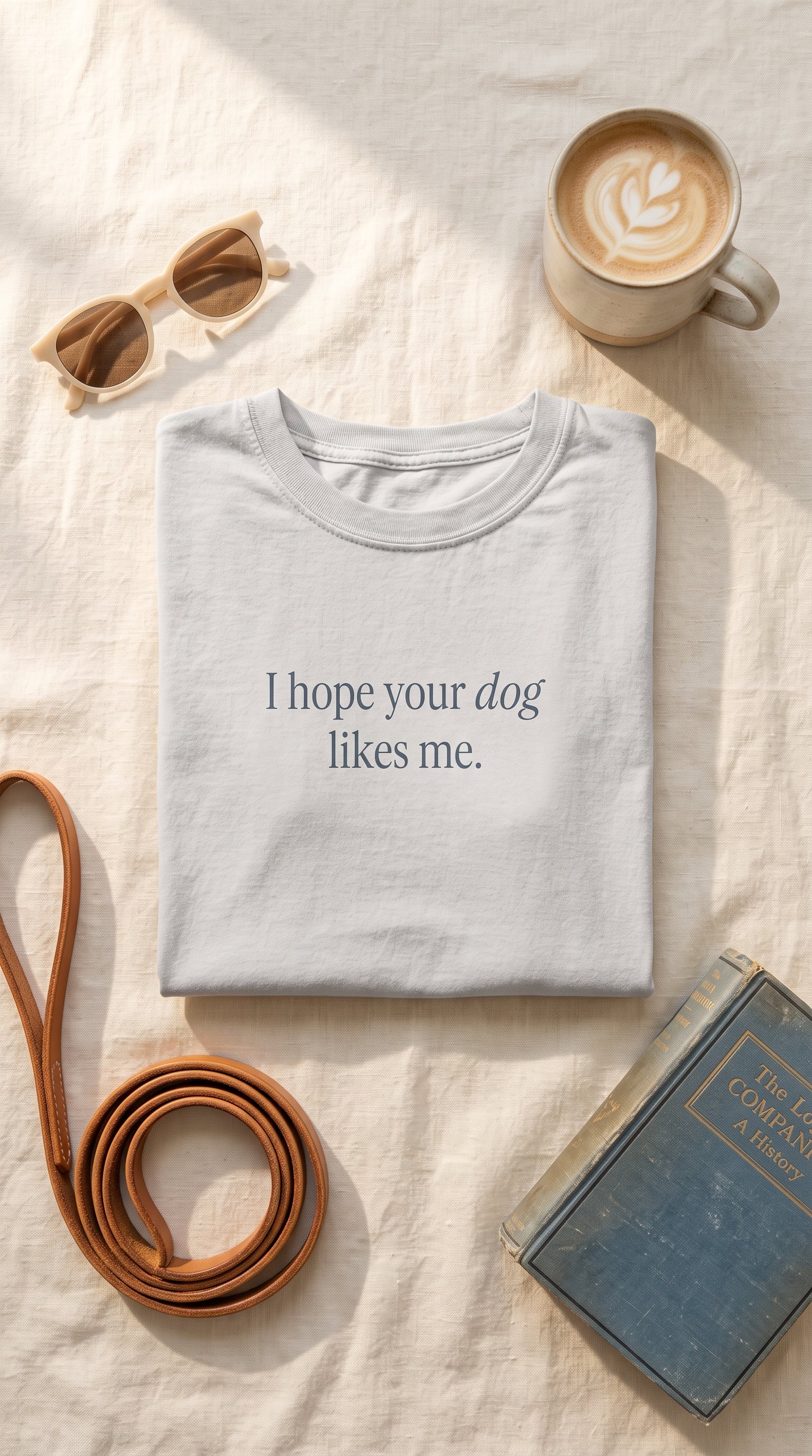 A folded light gray T-shirt with the printed phrase 'I hope your dog likes me.' in dark gray text, styled in a flat lay on a cream fabric surface with a coffee cup, sunglasses, a brown strap, and a blue book nearby.