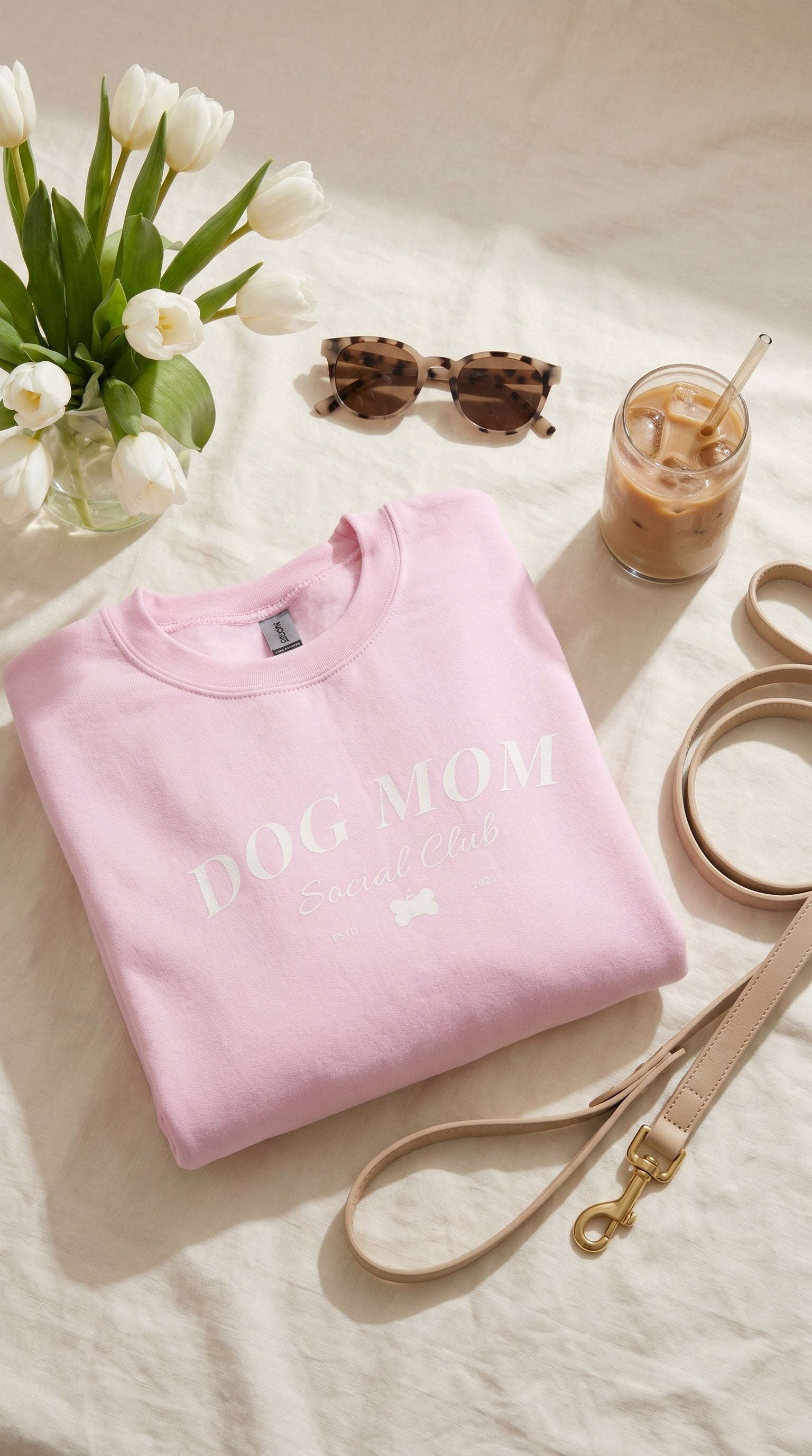 Pink sweatshirt with 'Dog Mom' text on a light surface with flowers, sunglasses, and a drink.