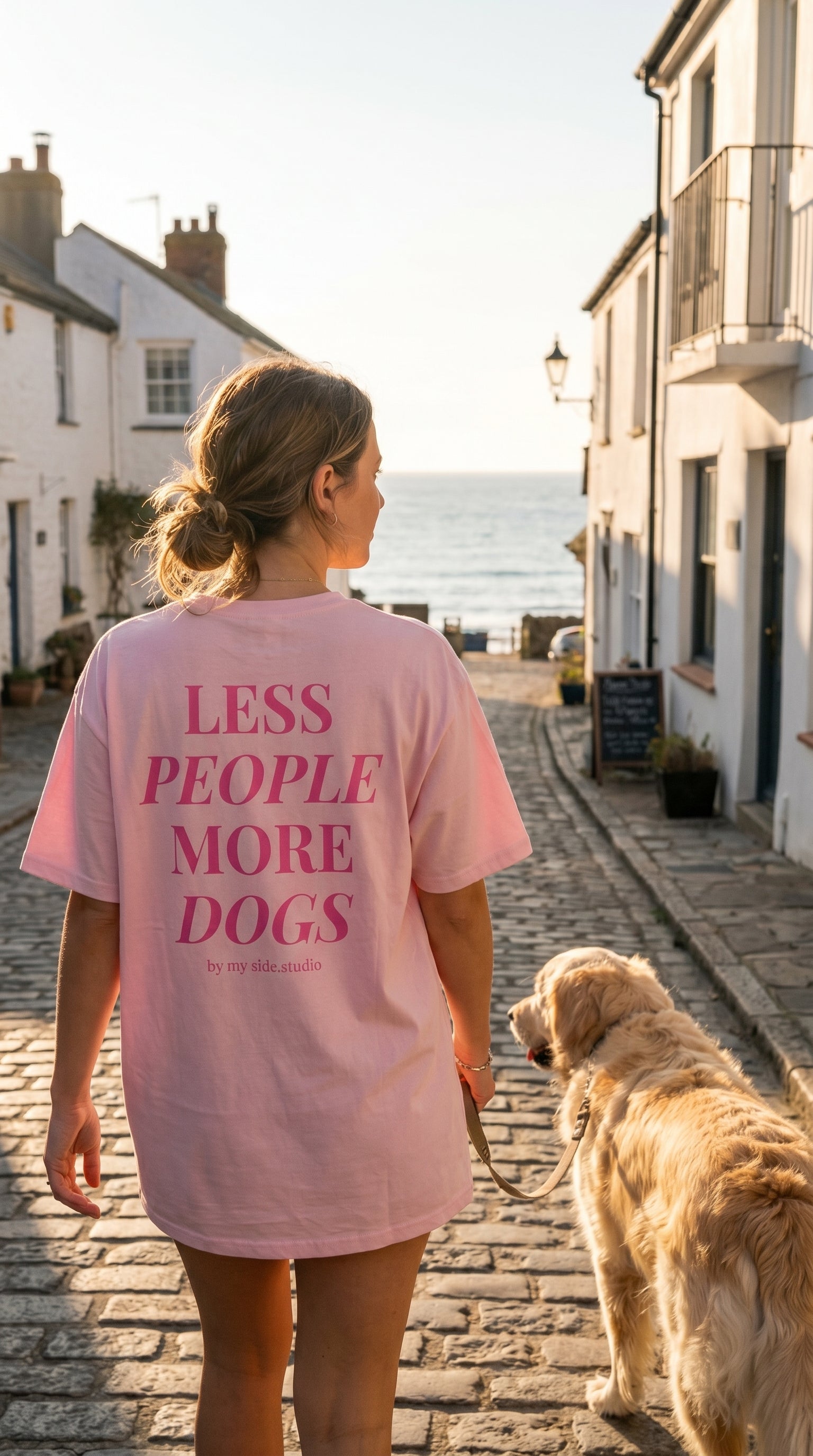 Woman walking a dog on a street with houses and ocean view, wearing a pink t-shirt with text.