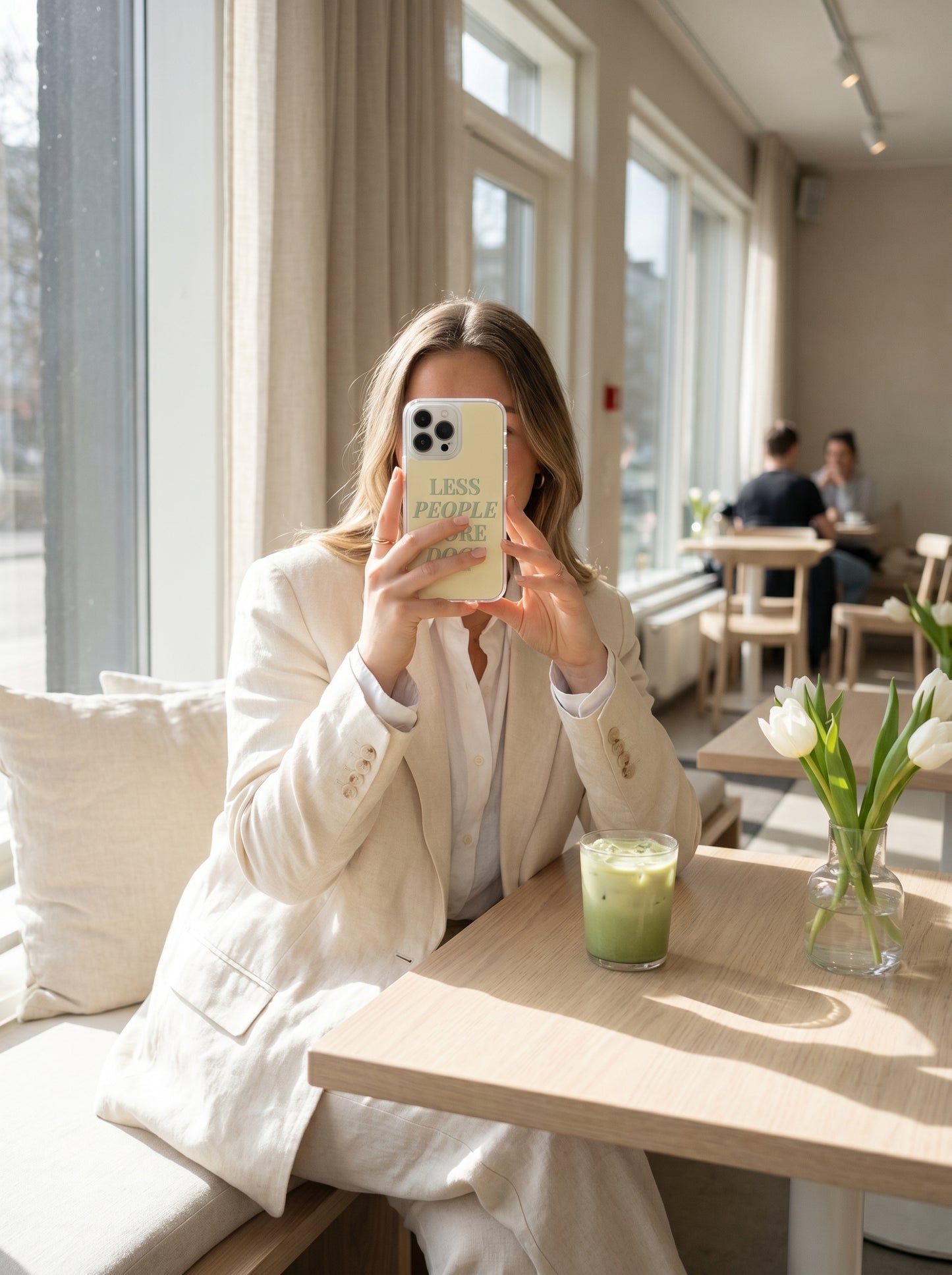 Woman holding a pastel yellow “Less People More Dogs” iPhone case beside an iced matcha latte in a bright Scandinavian-style coffee shop.