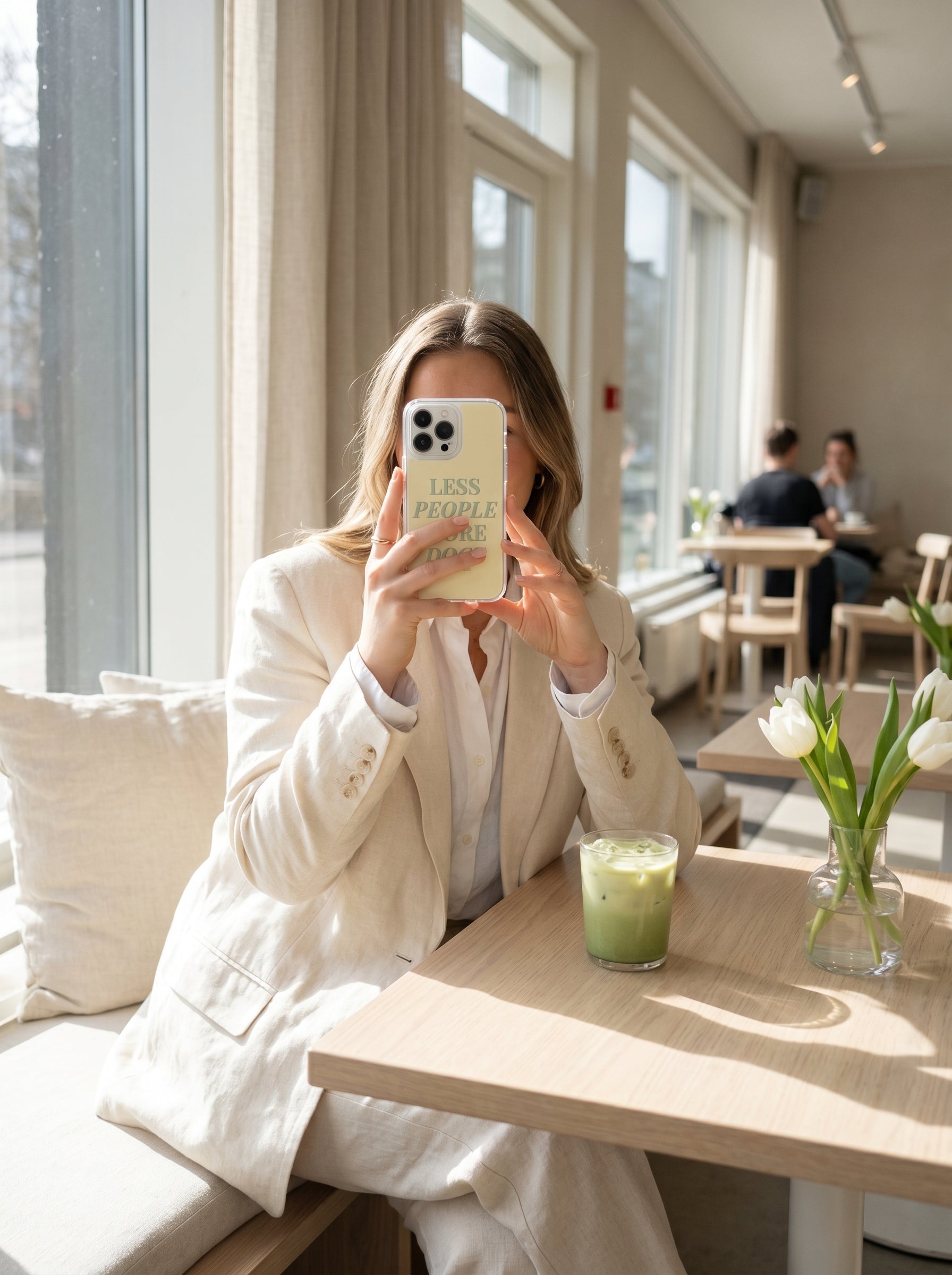 Woman holding a pastel yellow “Less People More Dogs” iPhone case beside an iced matcha latte in a bright Scandinavian-style coffee shop.