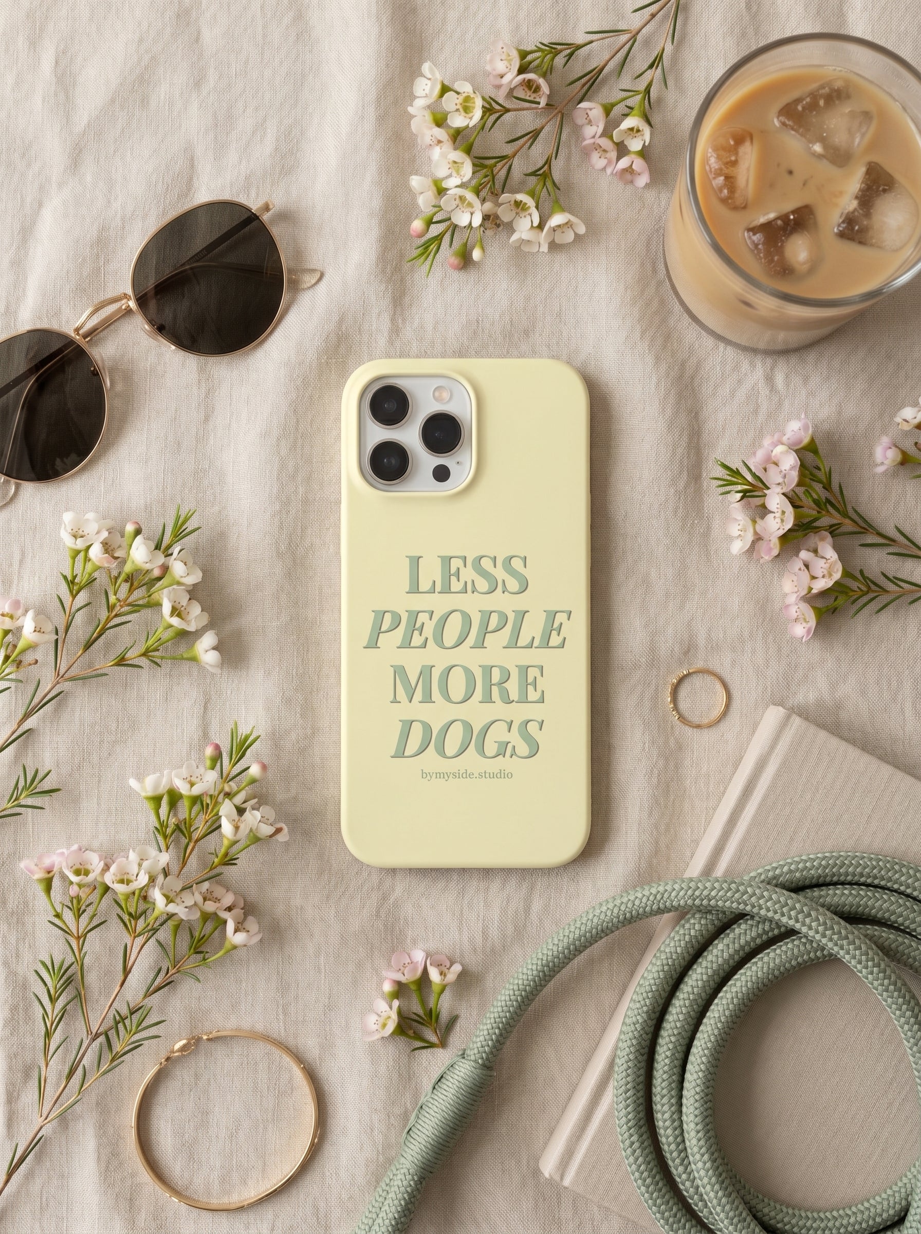 Minimal flat lay of the “Less People More Dogs” iPhone case styled with spring flowers, coffee, sunglasses, and neutral accessories.