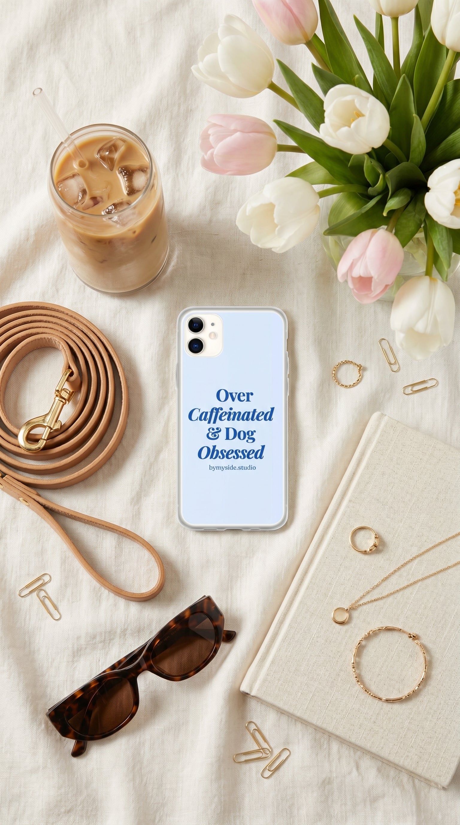 Flat lay of a pastel blue “Over Caffeinated & Dog Obsessed” iPhone case styled with coffee, flowers, and neutral accessories.
