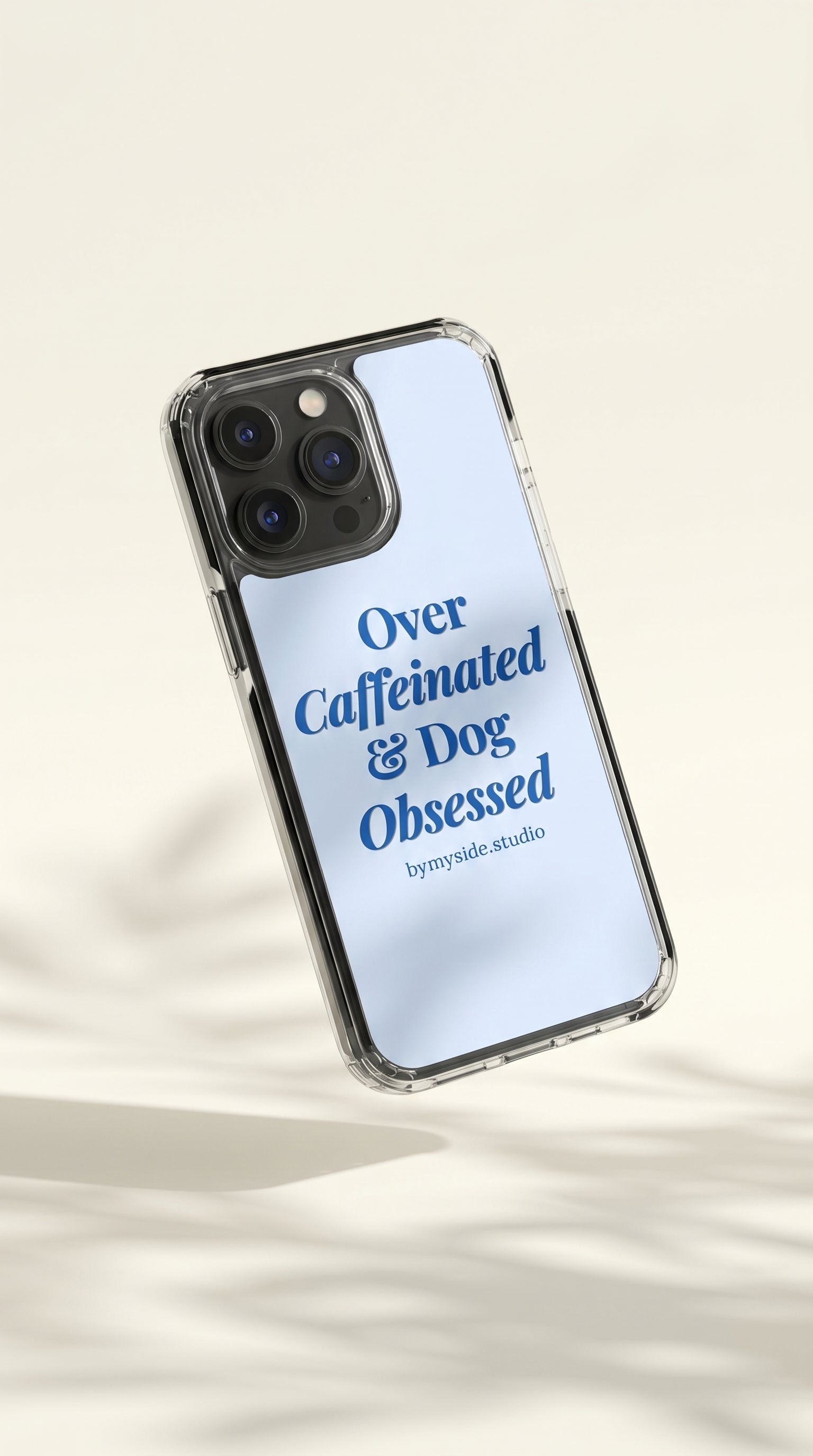 Minimal floating mockup of a pastel blue “Over Caffeinated & Dog Obsessed” iPhone case on a soft cream background.