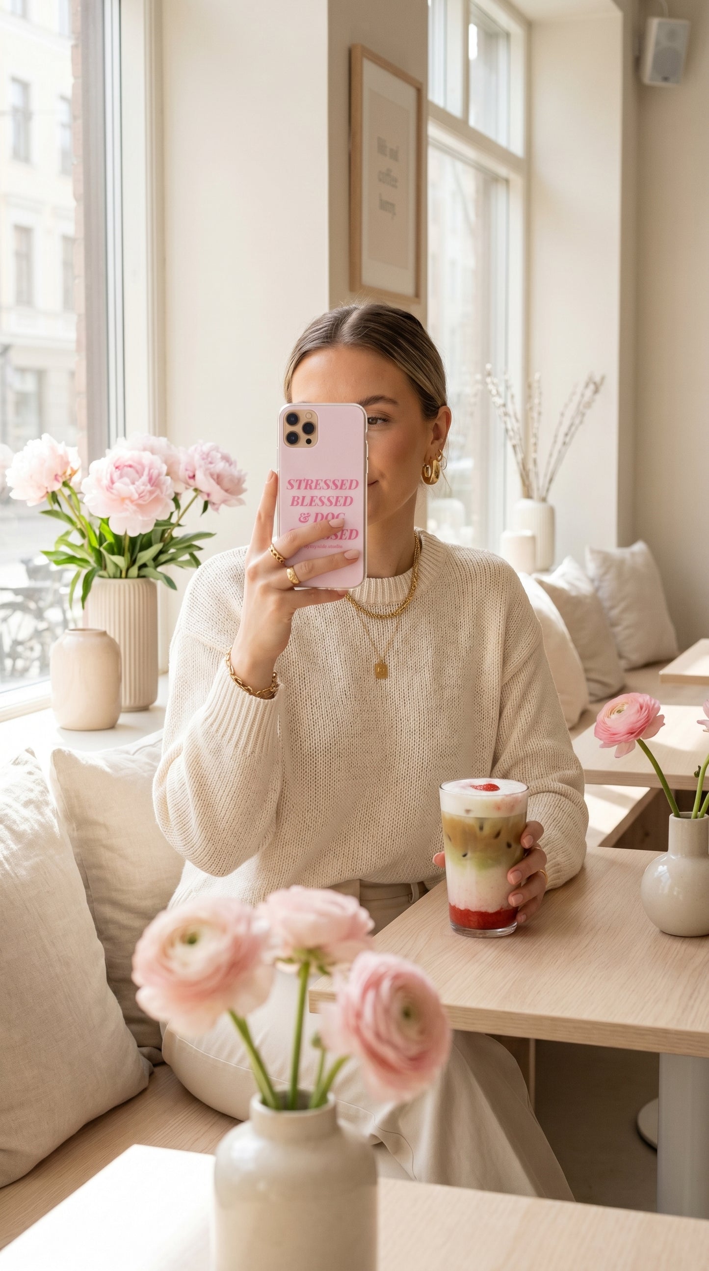 Woman holding a pastel pink “Stressed Blessed & Dog Obsessed” iPhone case beside an iced matcha latte in a bright spring café.