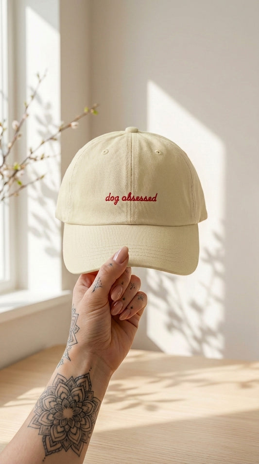 Beige cap with 'dog obsessed' text held by a hand with tattoos, against a bright window background.