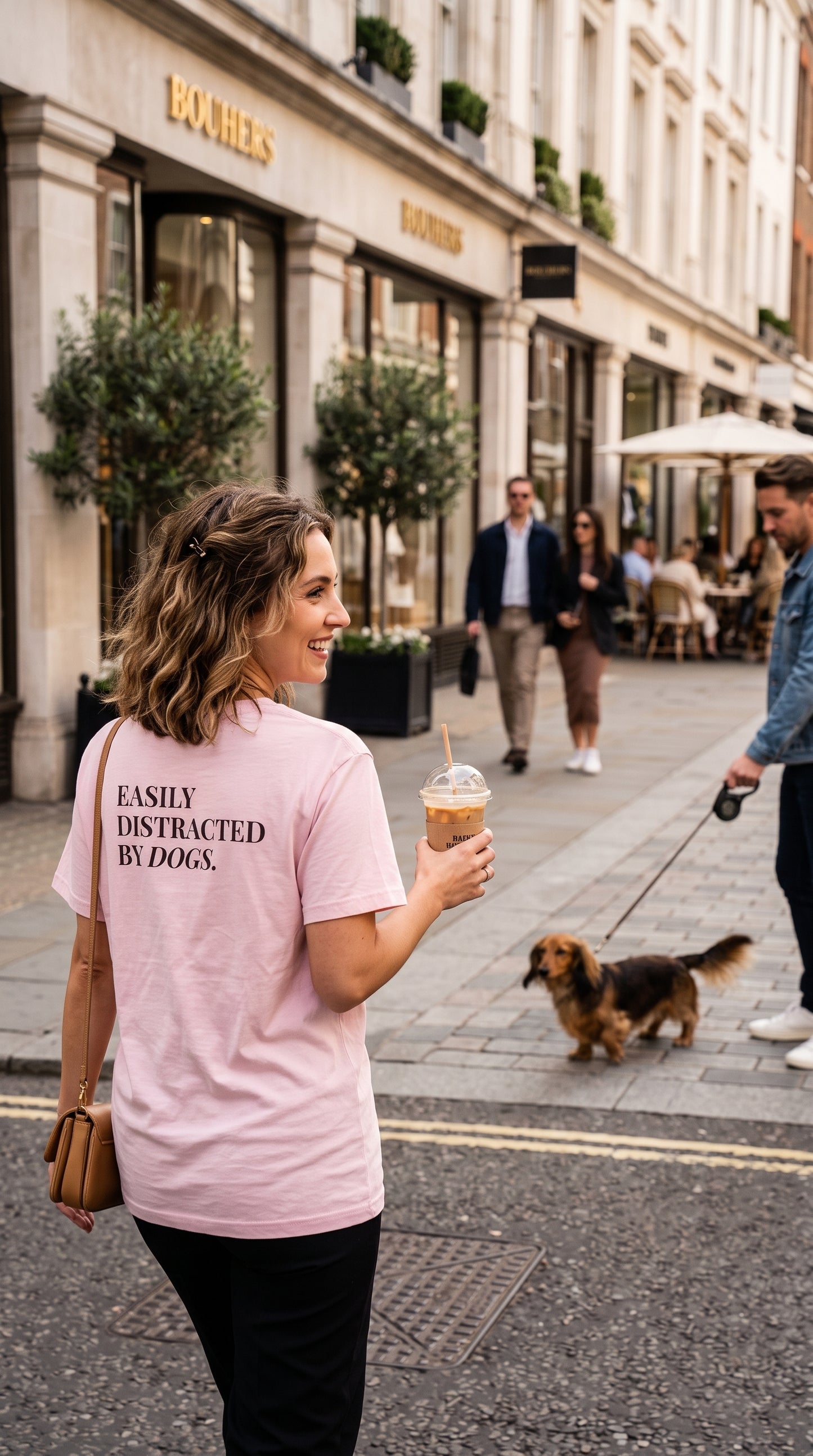 A woman walking on a city street wearing a light pink t-shirt with the text 'EASILY DISTRACTED BY DOGS.' printed on the back.