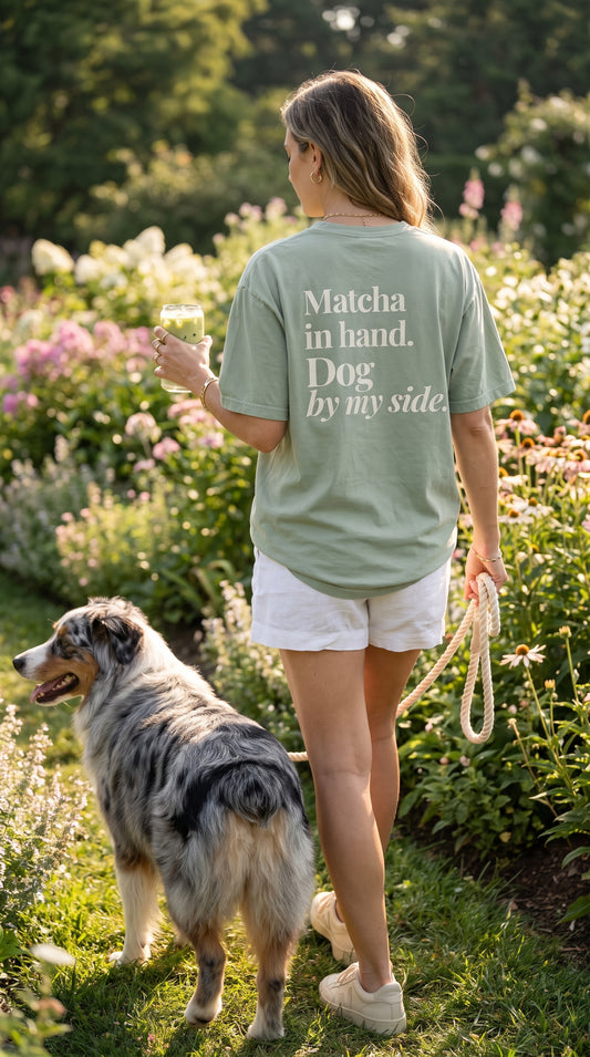 A woman wearing a sage green graphic t-shirt and white shorts walks with her dog in a sunny garden. The back of the t-shirt features the white text 'Matcha in hand.