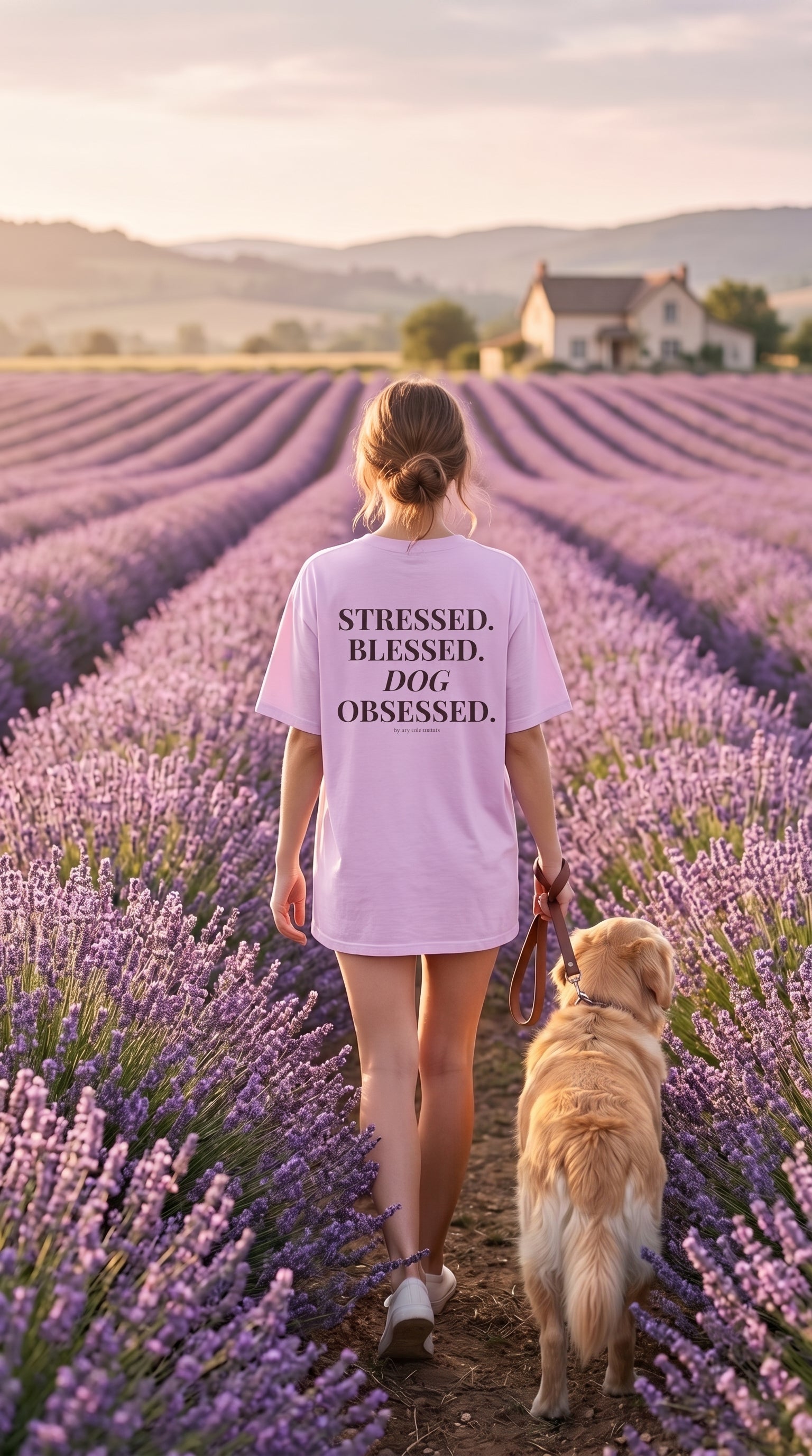 A woman and her golden retriever dog walk through a lavender field at sunset, viewed from behind. The woman wears a light pink oversized T-shirt with the black printed text 'STRESSED, BLESSED, DOG OBSESSED.