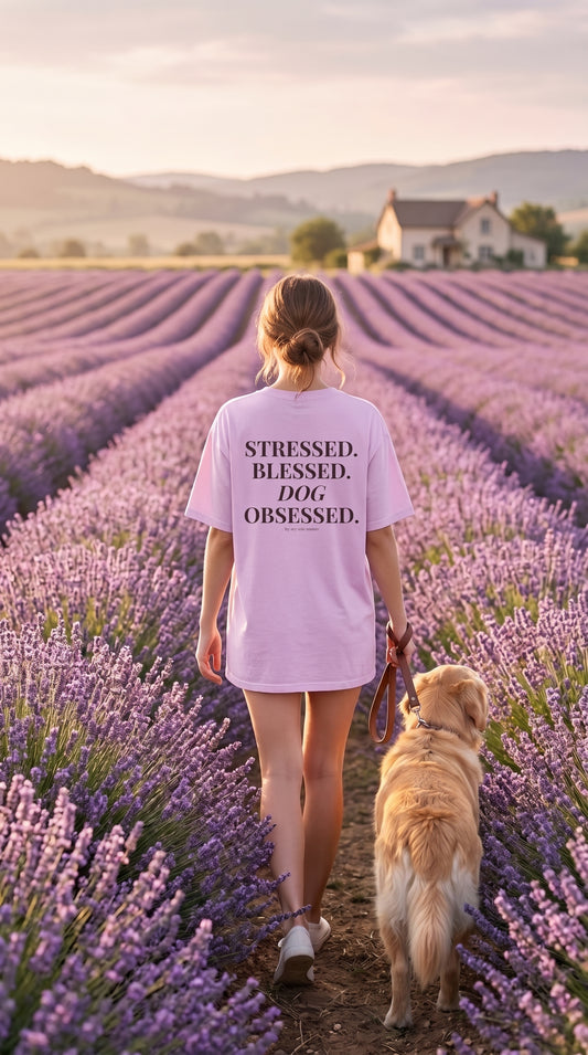 A woman and her golden retriever dog walk through a lavender field at sunset, viewed from behind. The woman wears a light pink oversized T-shirt with the black printed text 'STRESSED, BLESSED, DOG OBSESSED.