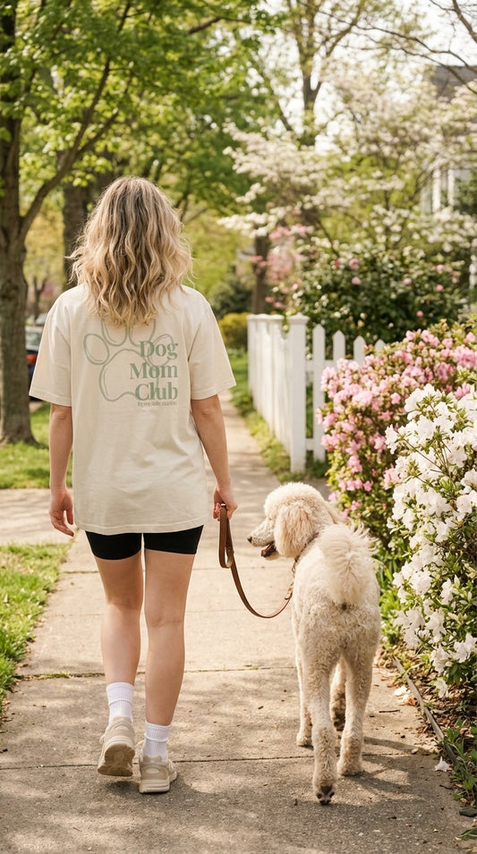 Woman walking a dog on a leash in a park-like setting with flowers and a white picket fence.