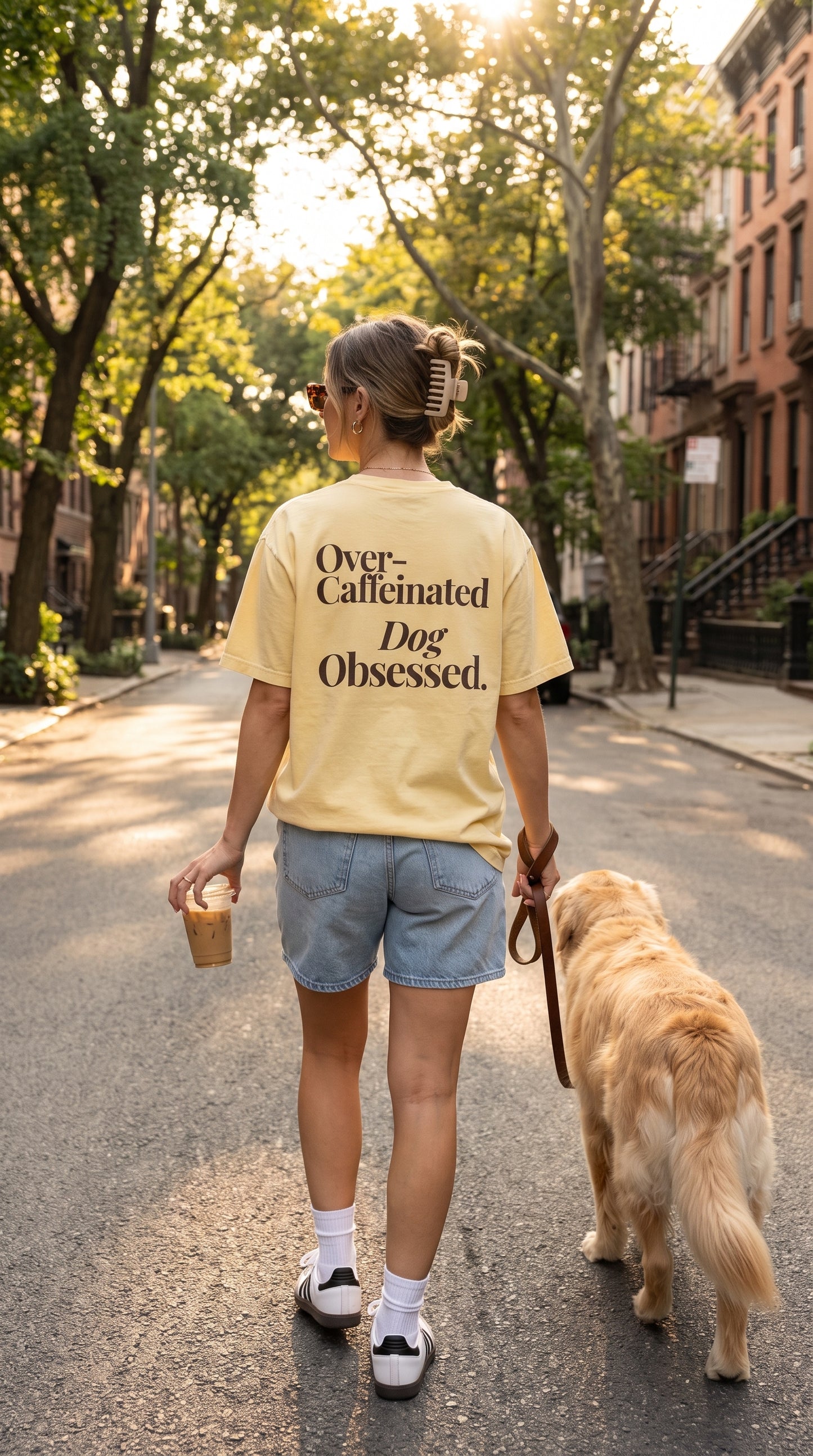 A woman seen from behind walking a golden retriever on a city street, wearing a pale yellow graphic T-shirt with the text 'Over-Caffeinated Dog Obsessed.' in black lettering, paired with denim shorts and white sneakers.