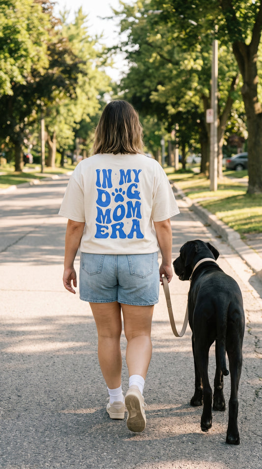 A woman walking a black dog on a paved path outdoors, viewed from behind. She is wearing a cream-colored graphic T-shirt with bold blue text that reads 'IN MY D&G MOM ERA' and light denim shorts.