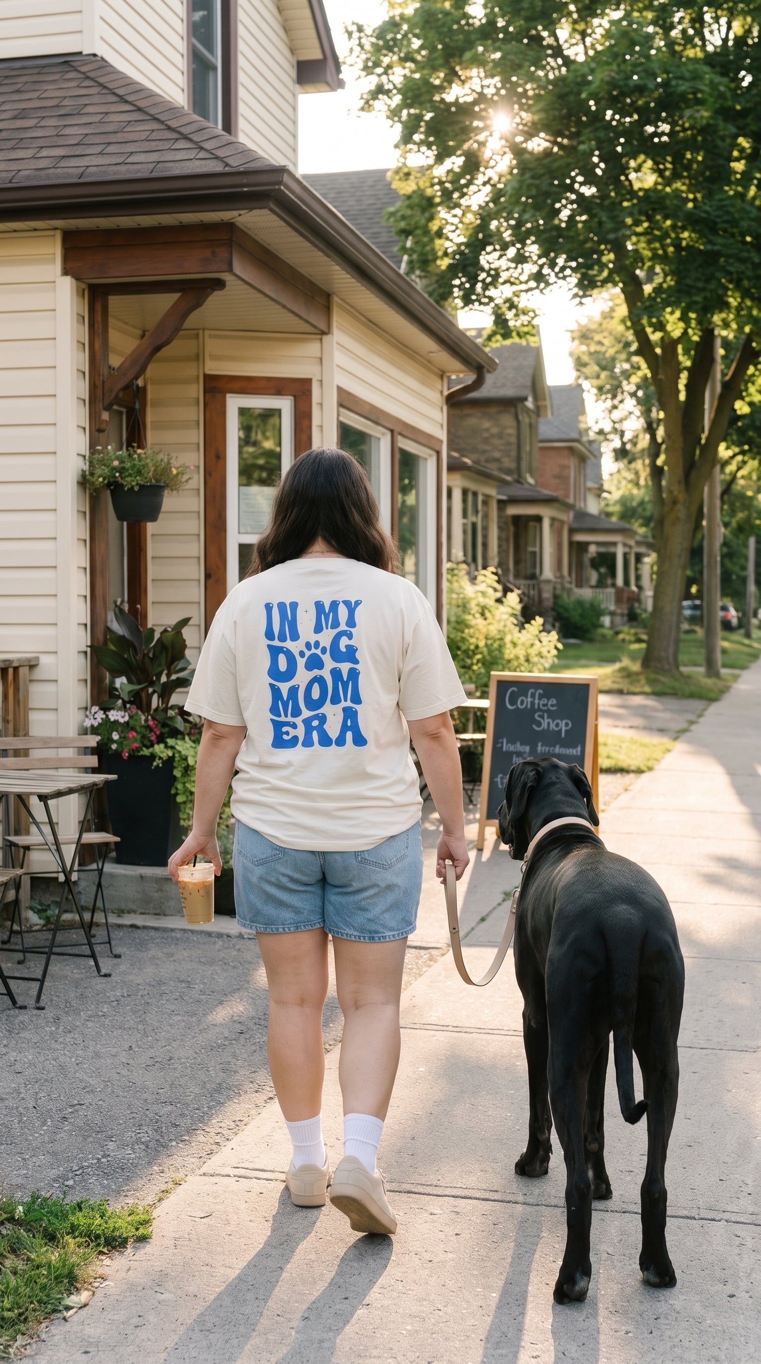 A woman walking a black dog on a sidewalk next to a house, wearing a cream t-shirt with blue text that reads 'IN MY DOG MOM ERA'.