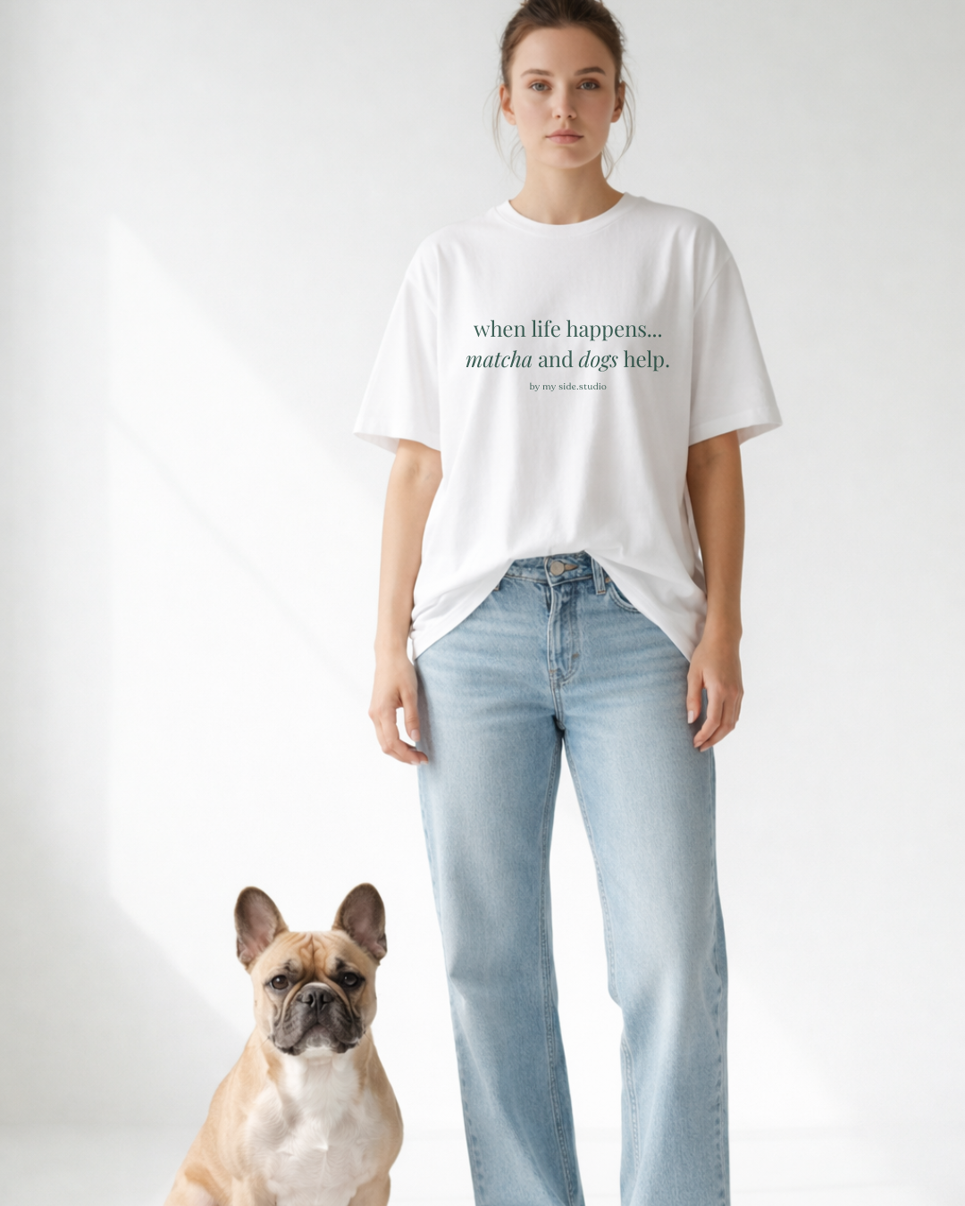 Person wearing a white t-shirt with text and light blue jeans standing next to a dog on a plain background